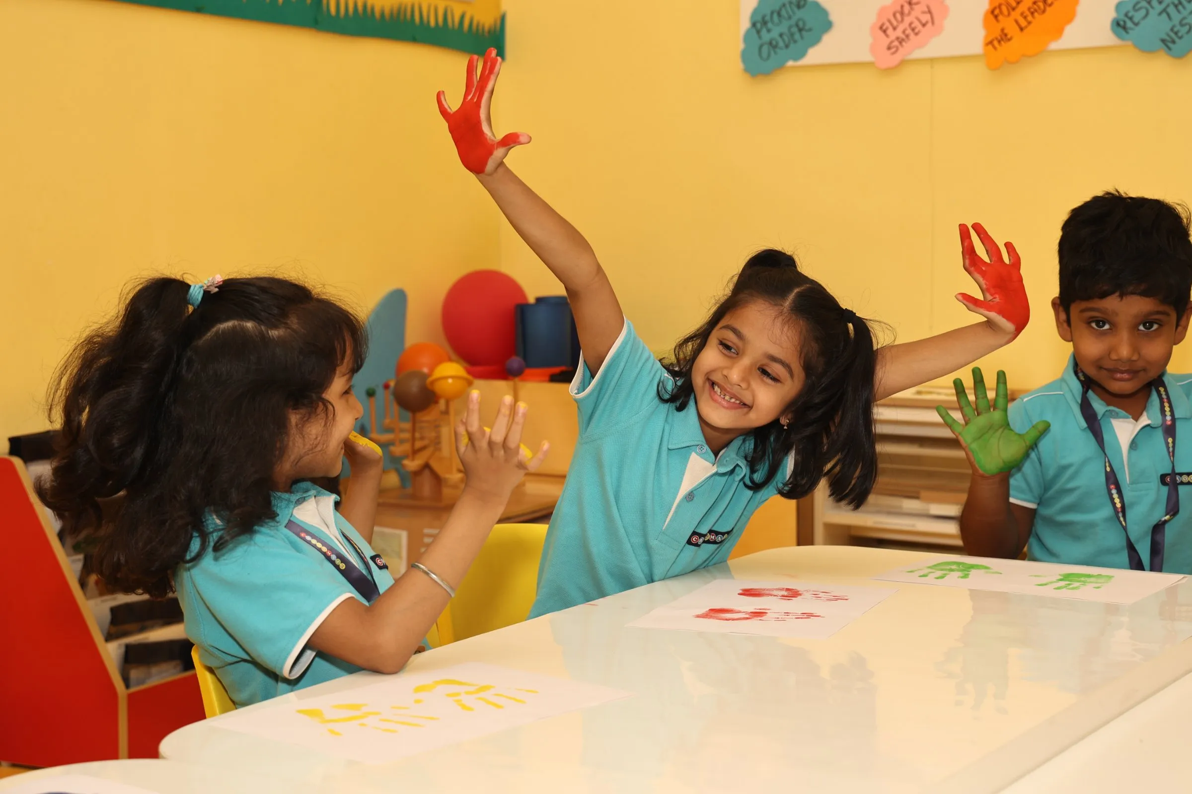 Children in their Crossmaze uniforms joyfully exploring colours and creating vibrant handprint art together in a bright, cheerful classroom.